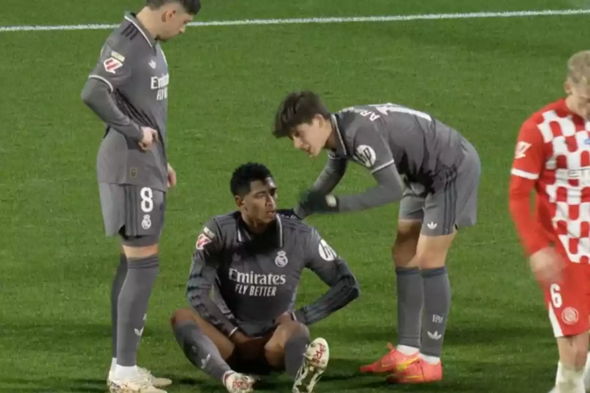 Real Madrid soccer players on the field, one of them sitting while the other two watch him.