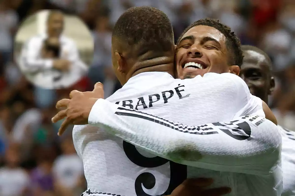 Two players hug each other celebrating a goal in a match, both wearing white uniforms and one of them has the name "Mbappé" on his back.