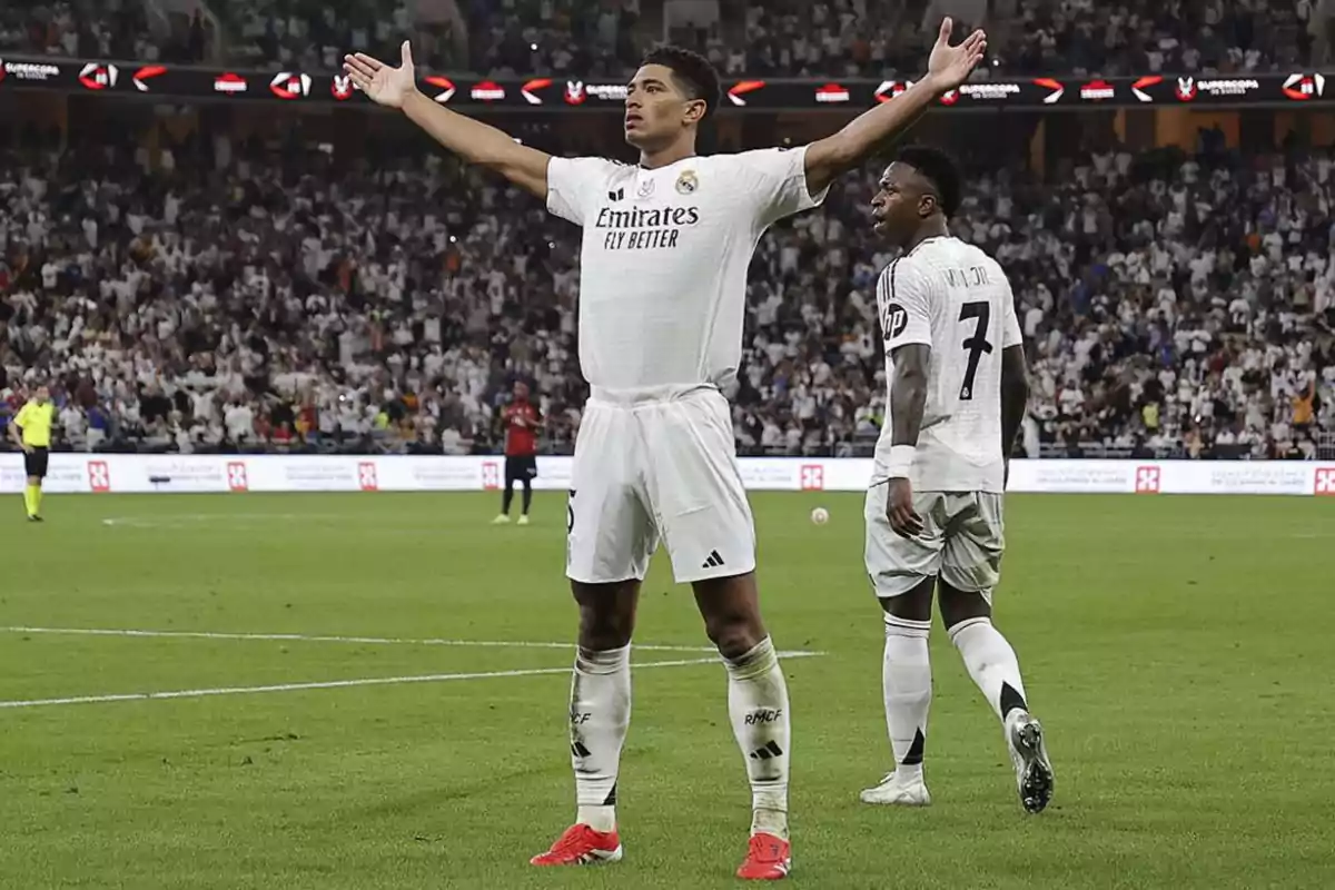 A soccer player with his arms outstretched celebrates on the field as another player walks near him, with a stadium full of spectators in the background. A soccer player with his arms outstretched celebrates on the field as another player walks near him, with a stadium full of spectators in the background.