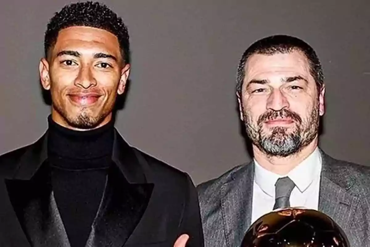 Dos hombres posando juntos, uno de ellos sostiene un trofeo dorado.