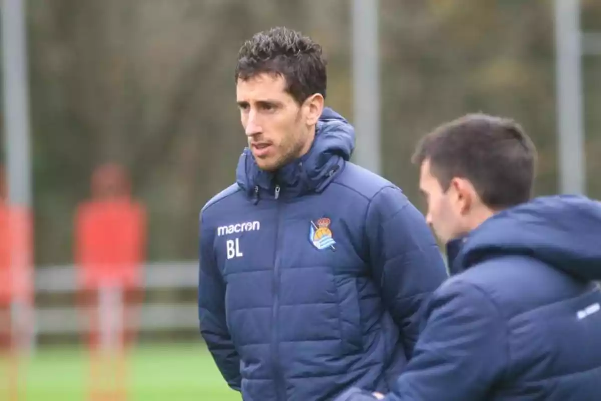 A man in a blue football team jacket is standing on a training field next to another person. A man in a blue football team jacket is standing on a training field next to another person.