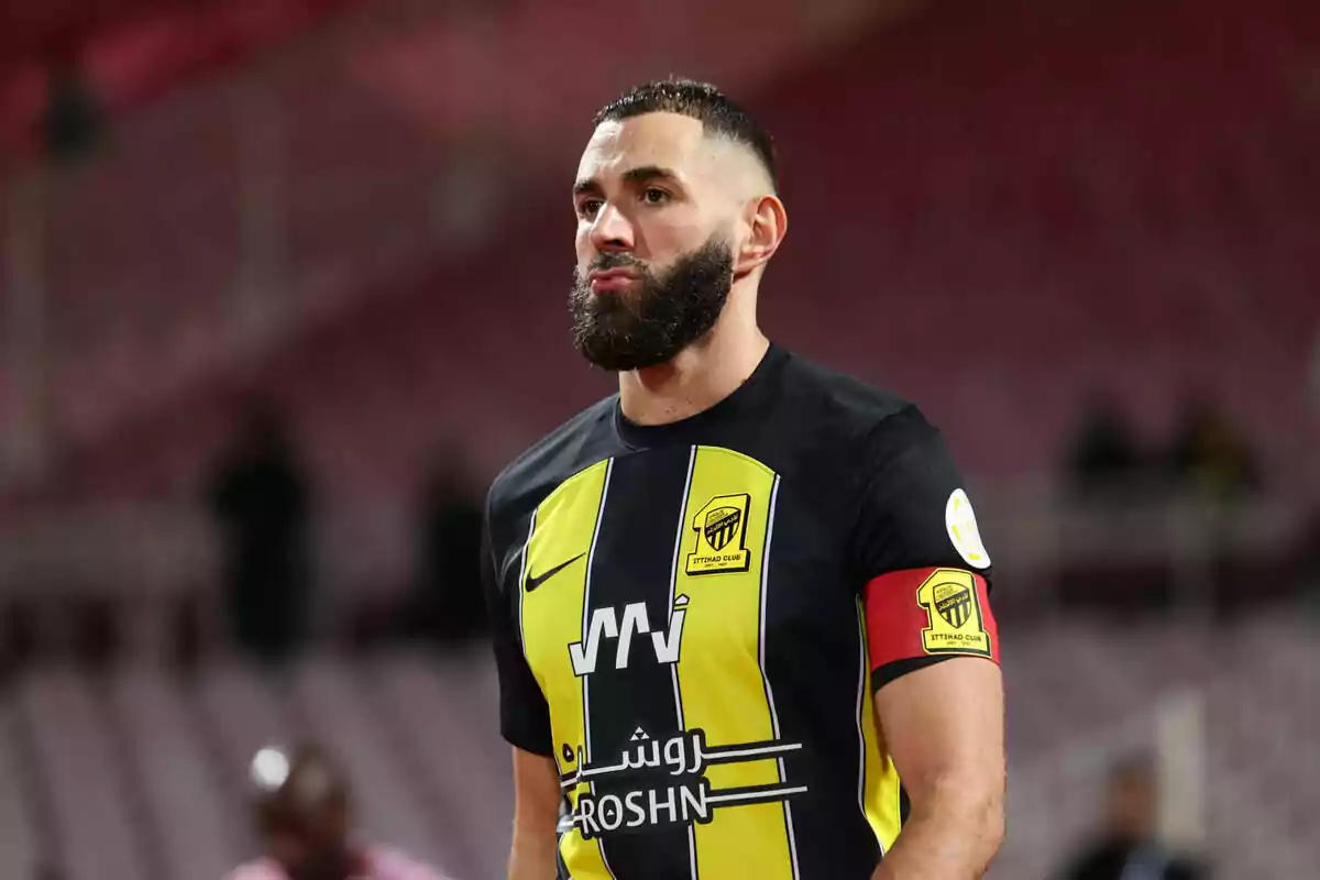 A soccer player with a beard and short hair wears a black and yellow jersey with a red captain's armband in a stadium. A soccer player with a beard and short hair wears a black and yellow jersey with a red captain's armband in a stadium.
