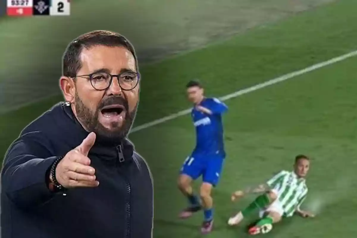 Un hombre con gafas y barba gesticula en primer plano mientras en el fondo se observa una jugada de fútbol con dos jugadores en el suelo.