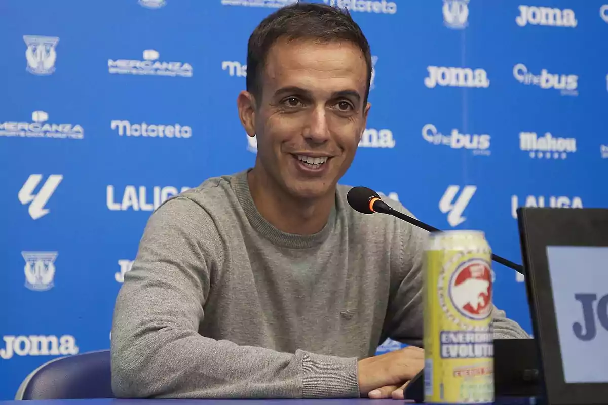 A smiley man is on the table at a press conference with a blue background full of logo and energy links.