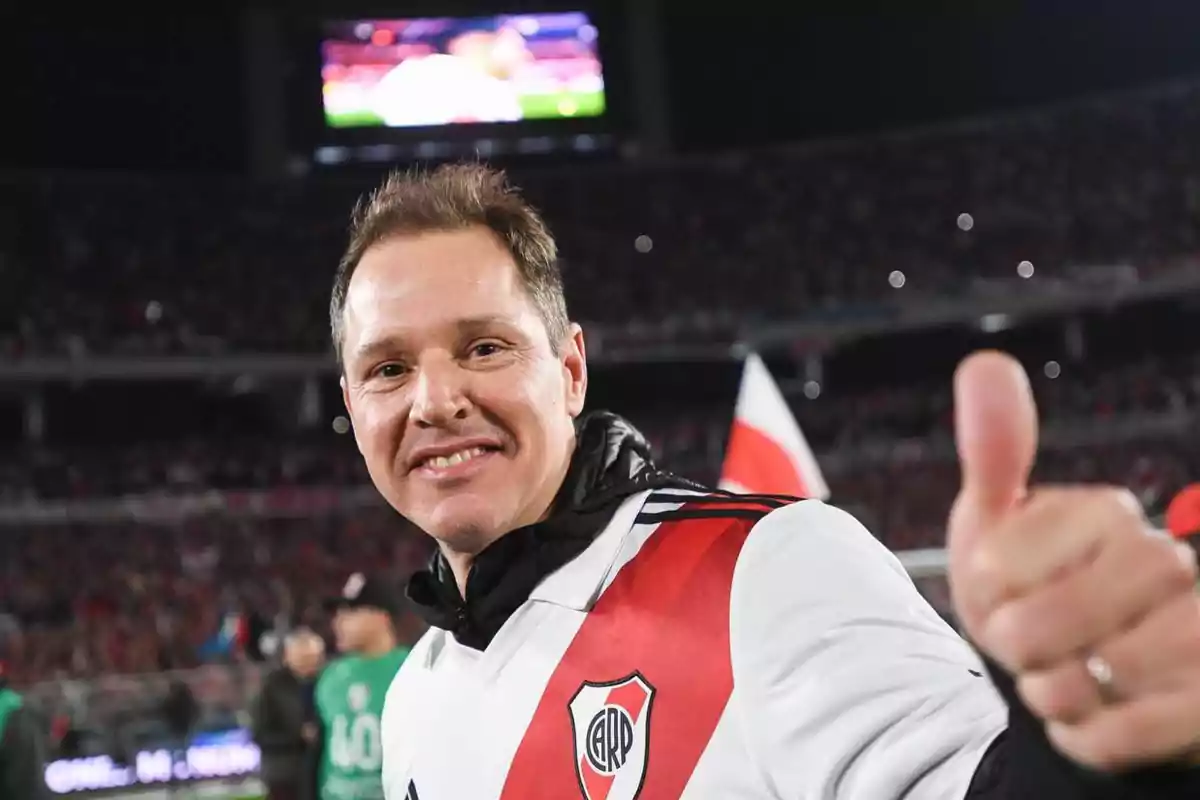 A smiling man wearing a River Plate jersey gives a thumbs-up gesture in a stadium full of spectators. A smiling man wearing a River Plate jersey gives a thumbs-up gesture in a stadium full of spectators.