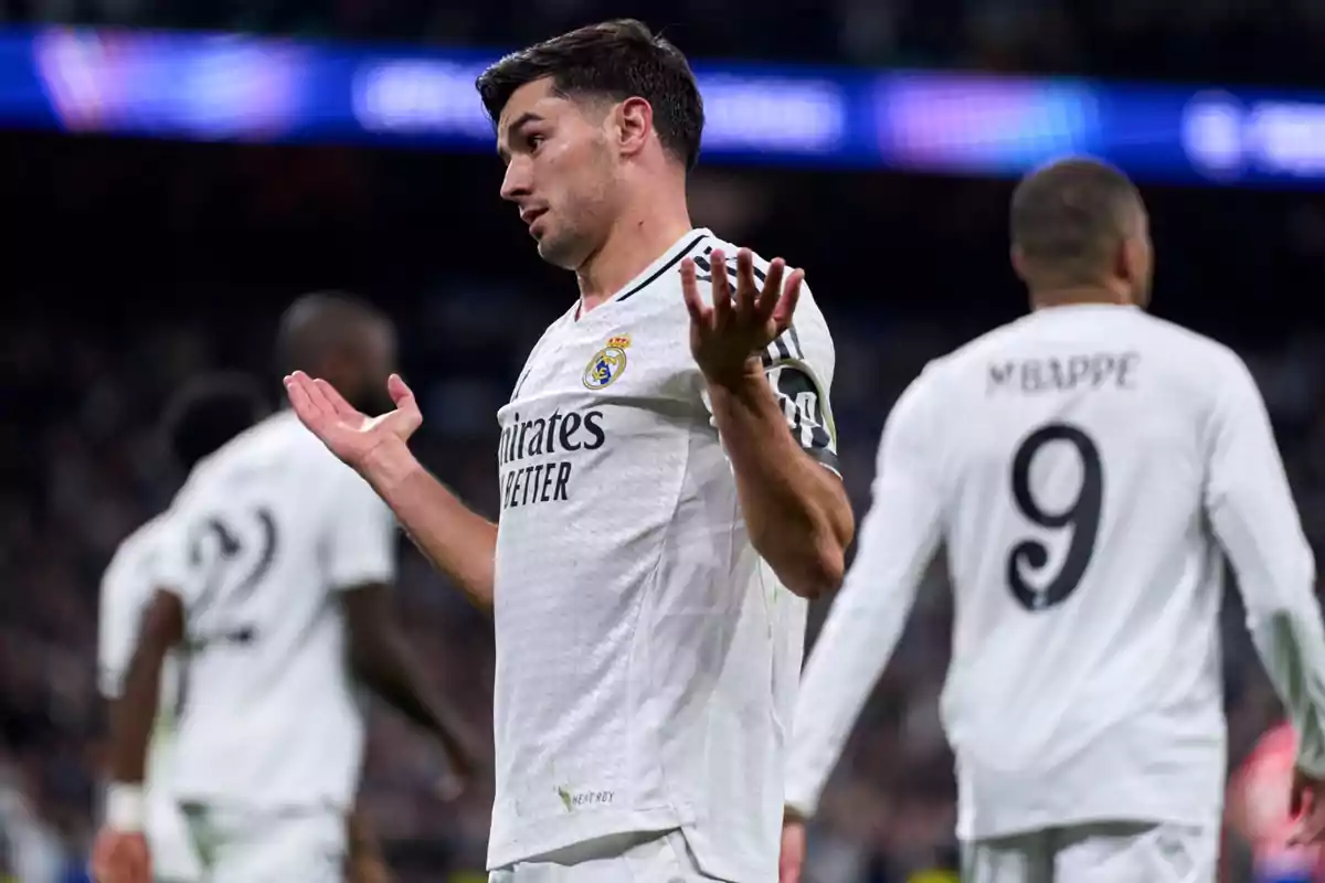 Un jugador de fútbol con el uniforme blanco del Real Madrid levanta las manos en un gesto de confusión o duda durante un partido.