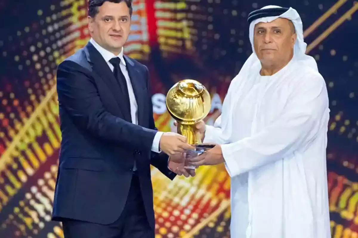 Two men on a stage hold a golden trophy together against a colorful background. Two men on a stage hold a golden trophy together against a colorful background.