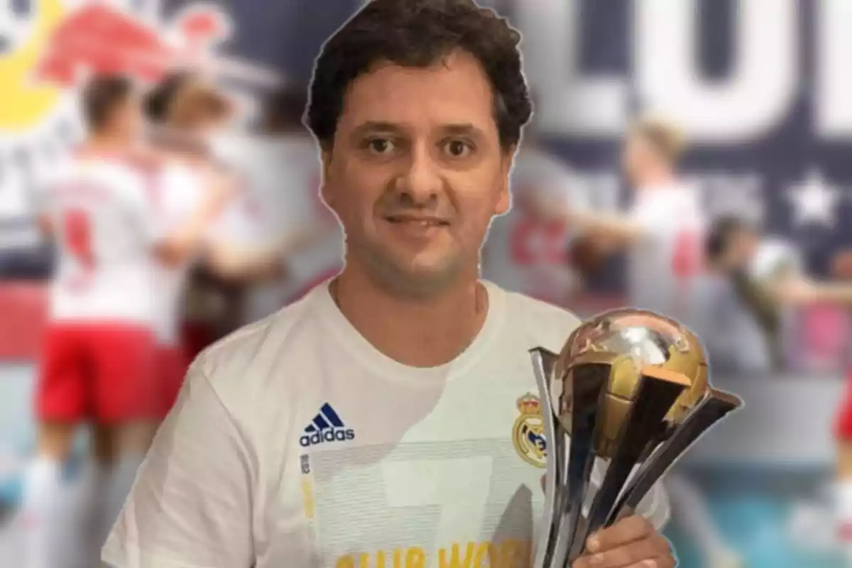 Un hombre con una camiseta del Real Madrid sostiene un trofeo frente a un fondo desenfocado de personas.