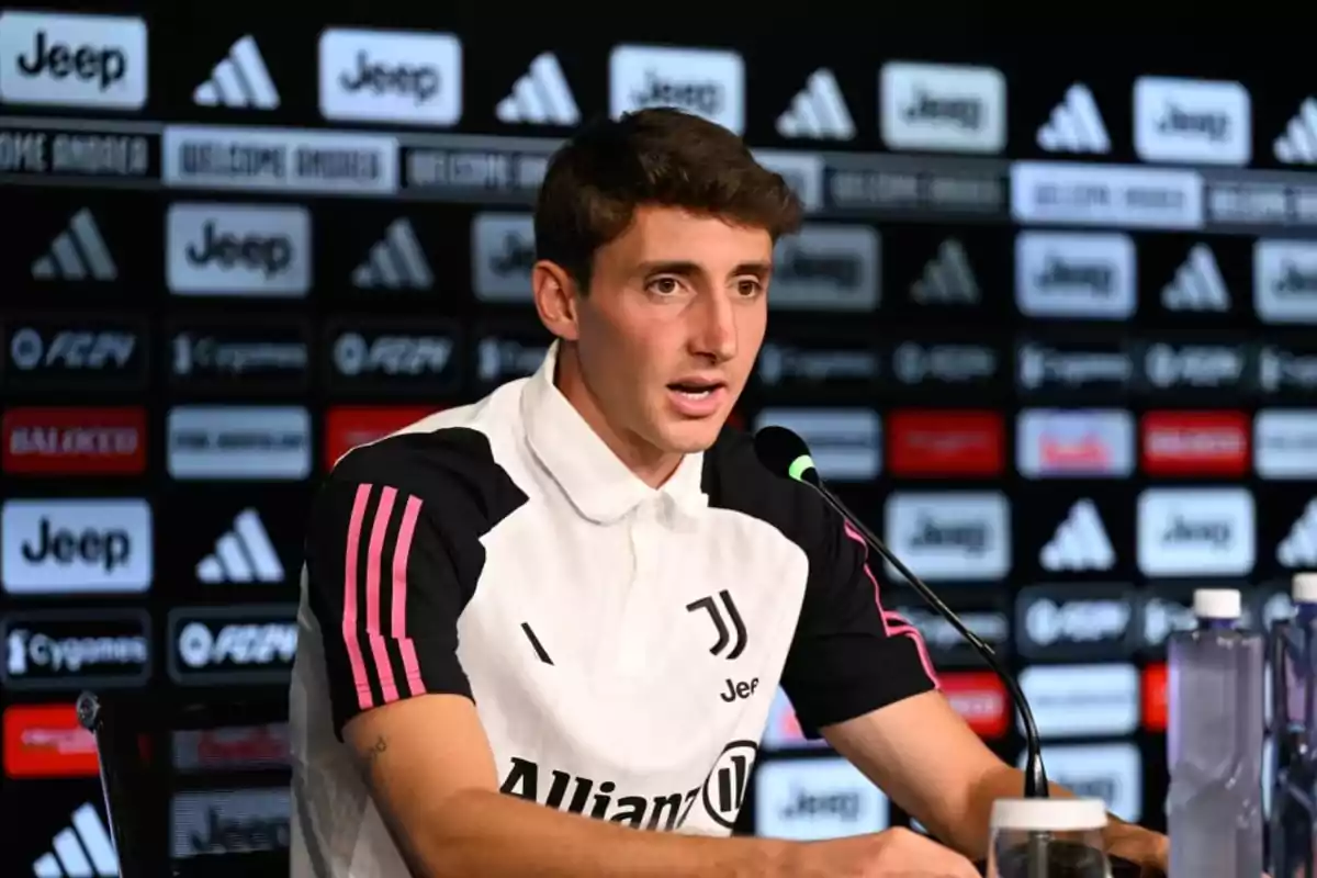 A soccer player at a press conference against a background of sponsor logos.