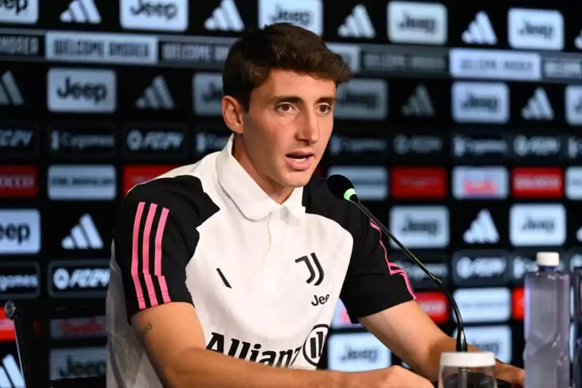 A soccer player at a press conference with a microphone in front of him and a background with sponsor logos. A soccer player at a press conference with a microphone in front of him and a background with sponsor logos.