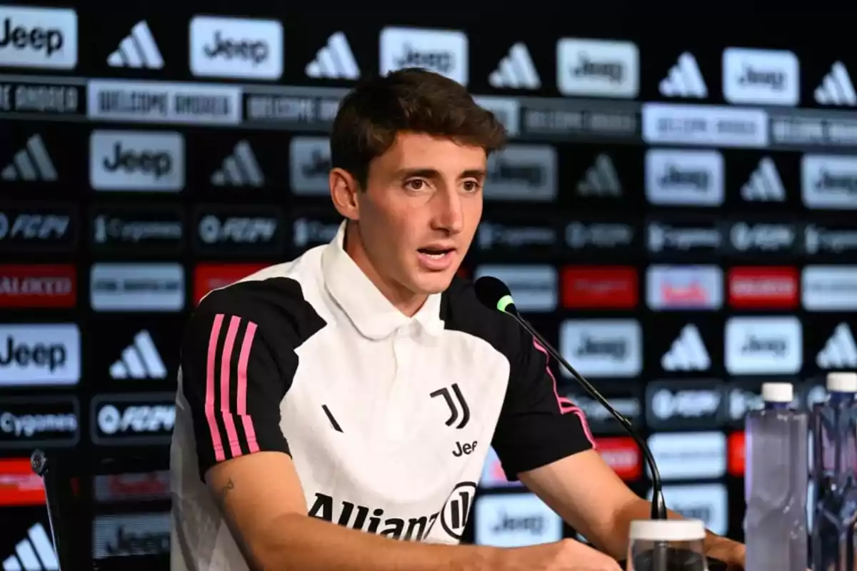 A soccer player at a press conference with a microphone in front of him and a background with sponsor logos.