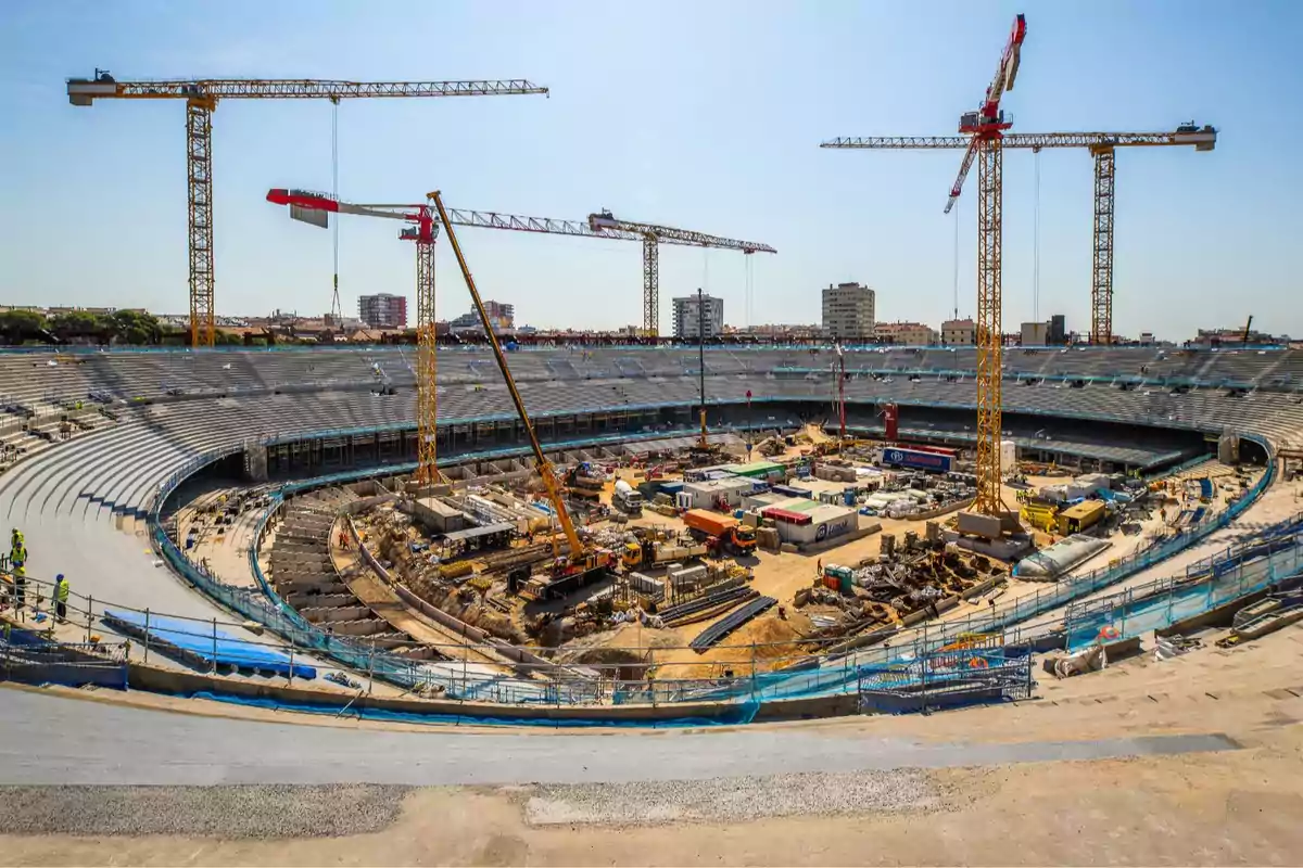 Construcción de un estadio con grúas y maquinaria pesada en el centro del campo y gradas parcialmente terminadas.