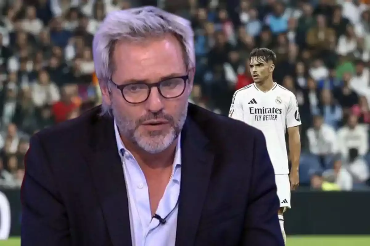 Un hombre con gafas y barba aparece en primer plano mientras un jugador de fútbol con uniforme blanco está en el fondo en un estadio lleno de espectadores.