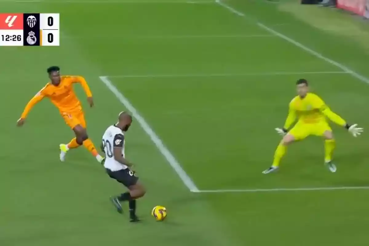 Un jugador de fútbol con uniforme blanco y negro se prepara para patear el balón mientras es perseguido por un jugador con uniforme naranja y el portero con uniforme amarillo se prepara para defender la portería.