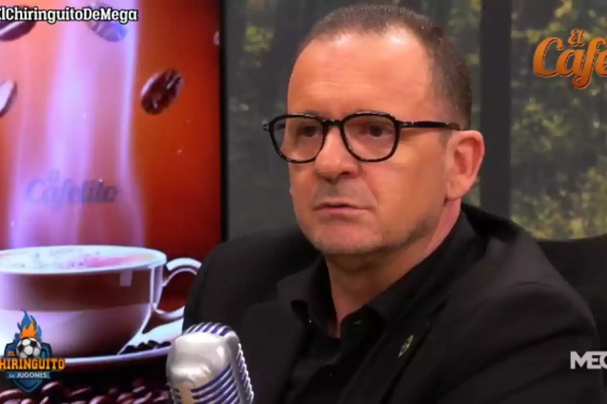 Un hombre con gafas y chaqueta oscura está sentado frente a un micrófono, con una pantalla detrás que muestra una imagen de una taza de café y granos de café.