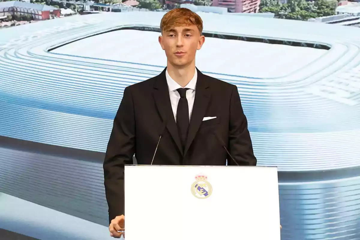 Joven con traje oscuro hablando en un podio con el escudo del Real Madrid y el estadio Santiago Bernabéu de fondo Joven con traje oscuro hablando en un podio con el escudo del Real Madrid y el estadio Santiago Bernabéu de fondo