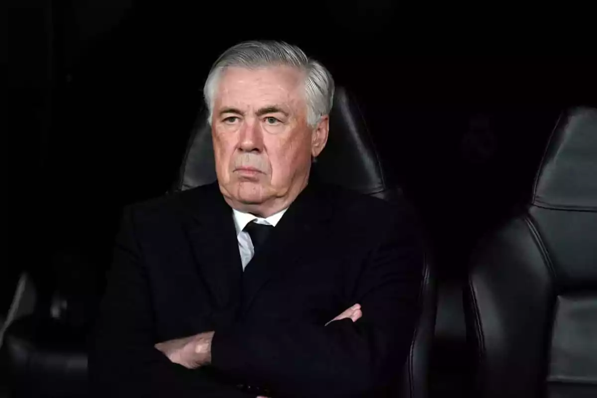 A gray-haired man in a dark suit is sitting with his arms crossed on a black leather seat, with a serious expression and a dark background. A gray-haired man in a dark suit is sitting with his arms crossed on a black leather seat, with a serious expression and a dark background.