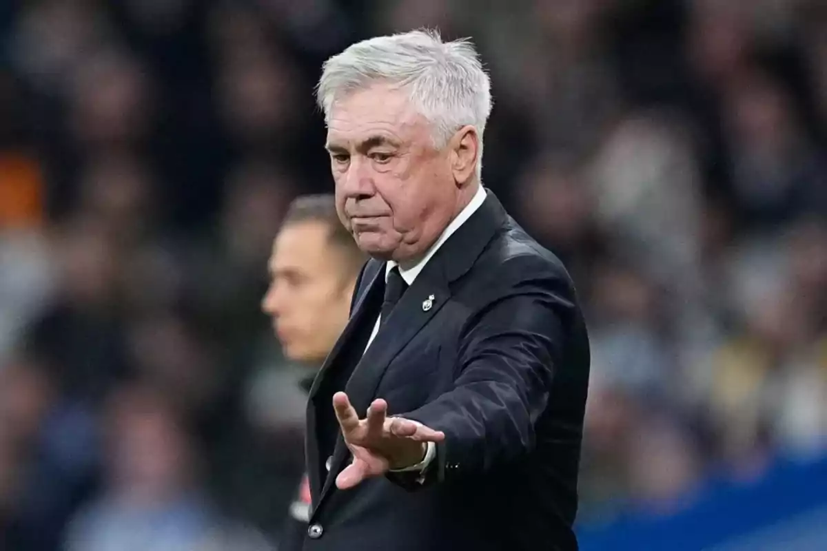 A gray-haired man in a dark suit gestures with his hand in a sports setting. A gray-haired man in a dark suit gestures with his hand in a sports setting.