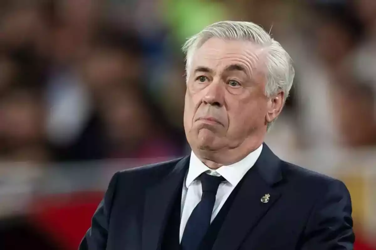 A gray-haired man with a pensive expression wears a dark suit with a tie, in a setting with a blurred background. A gray-haired man with a pensive expression wears a dark suit with a tie, in a setting with a blurred background.