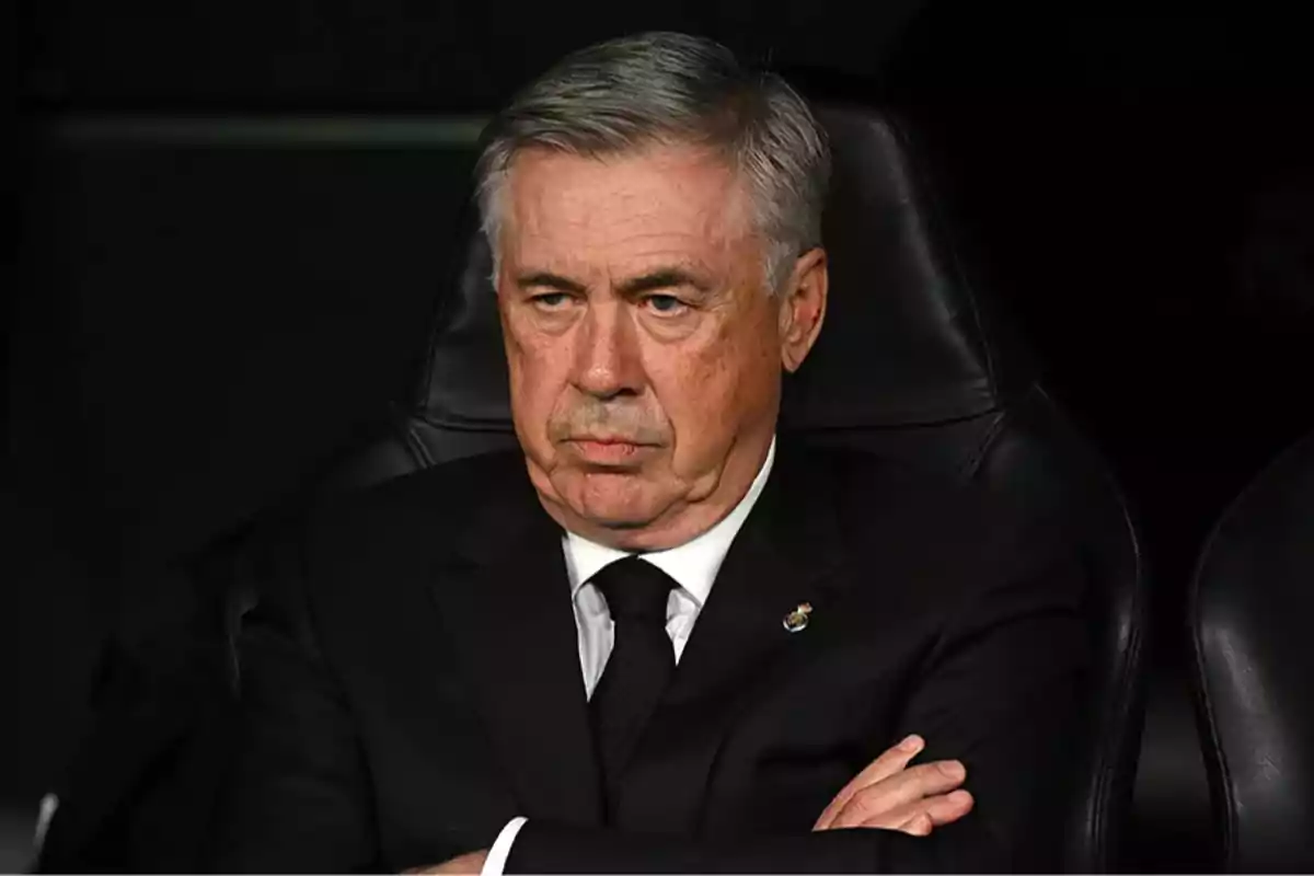 A man with gray hair and a serious expression sits with his arms crossed, wearing a dark suit. A man with gray hair and a serious expression sits with his arms crossed, wearing a dark suit.