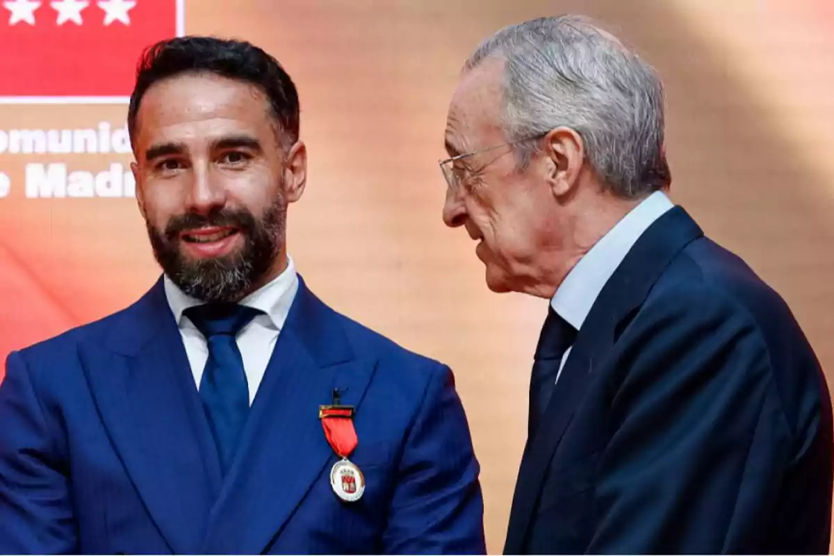 Dos hombres con traje conversan en un evento formal uno de ellos lleva una medalla roja en la solapa