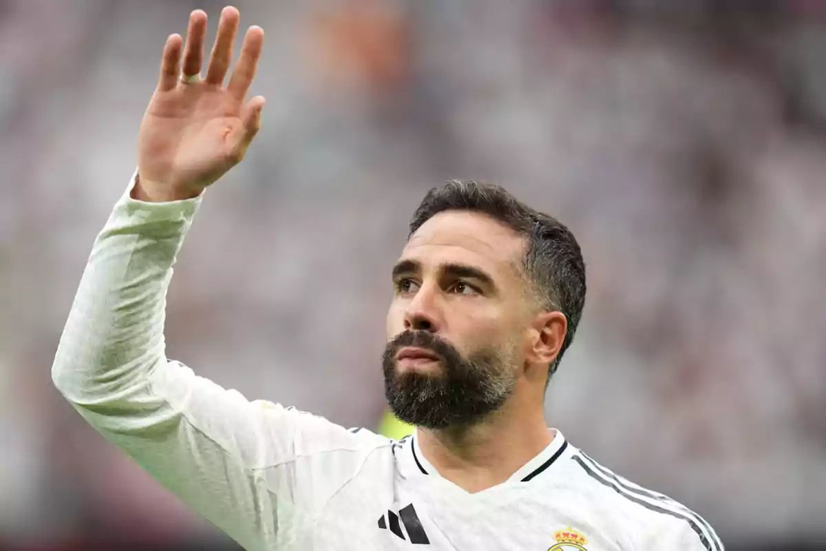 Un jugador de fútbol con barba y cabello corto levanta la mano mientras viste un uniforme blanco en un estadio.