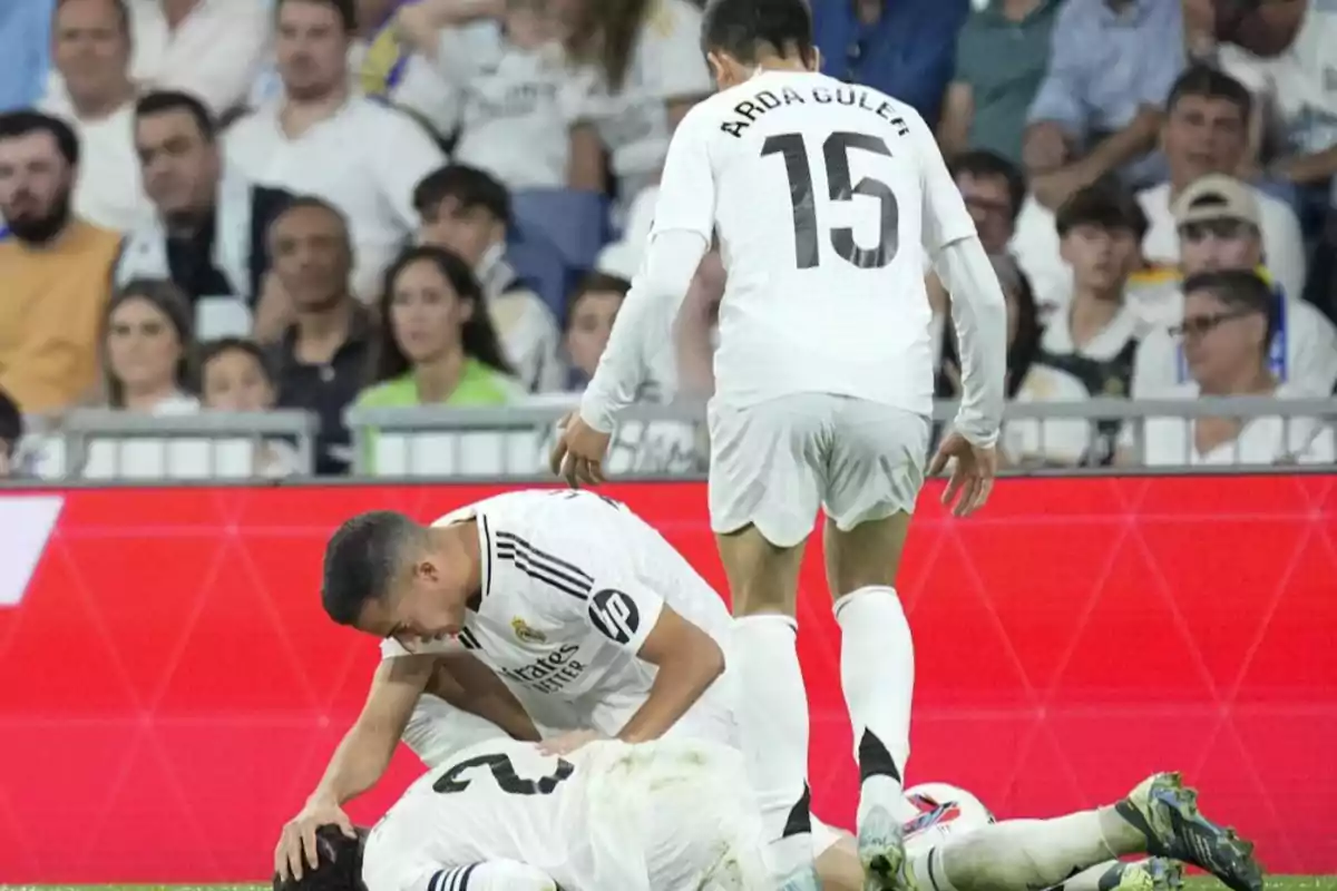 Real Madrid soccer players on the field, one of them on the ground while another assists him, with spectators in the background. Real Madrid soccer players on the field, one of them on the ground while another assists him, with spectators in the background.
