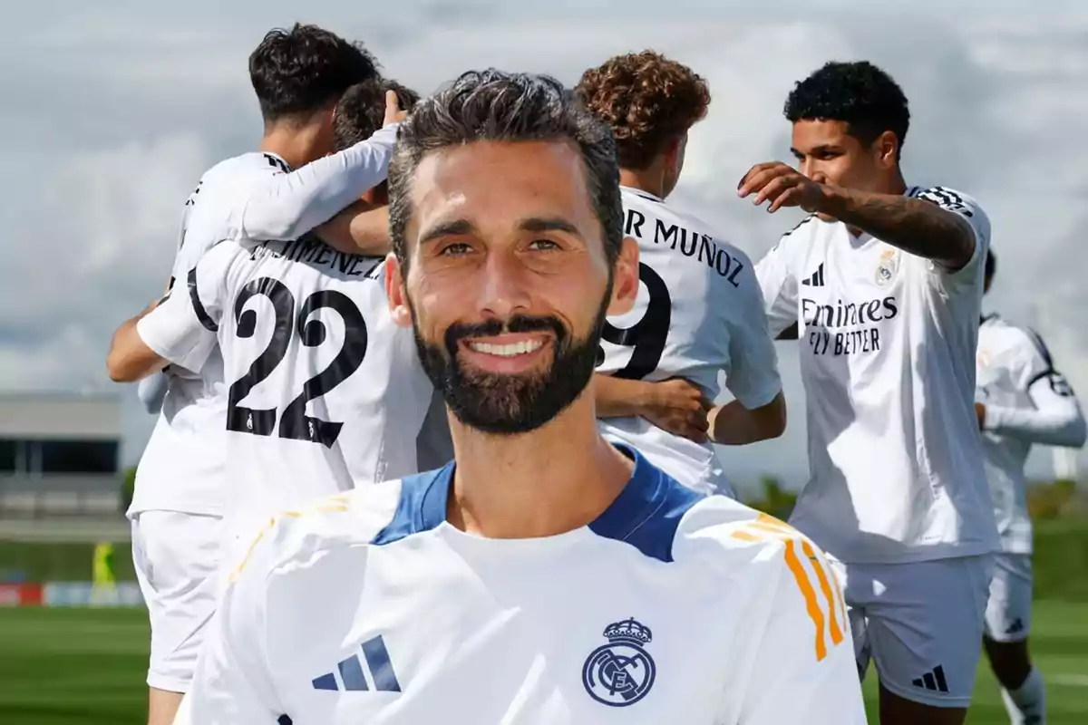 Un hombre con barba y camiseta del Real Madrid sonríe al frente mientras varios jugadores del mismo equipo celebran abrazados en el campo de fútbol