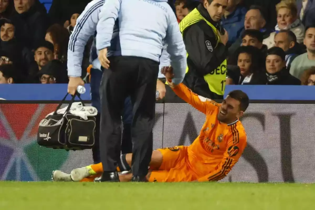 A player dressed in orange is on the ground with a pained expression while being assisted by medical staff on the field, surrounded by spectators.