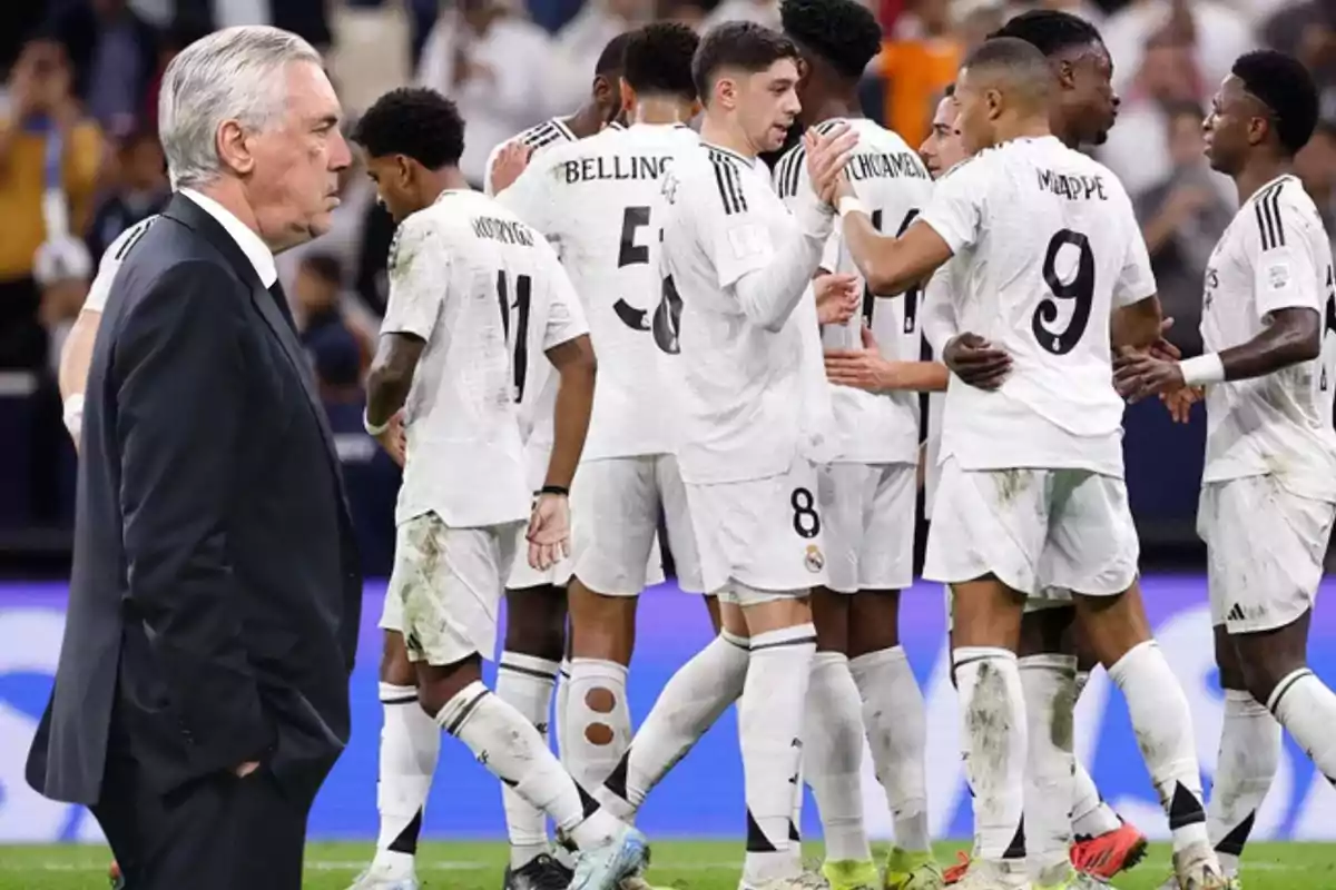 Un entrenador observa a un grupo de jugadores de fútbol vestidos con uniformes blancos mientras celebran en el campo.