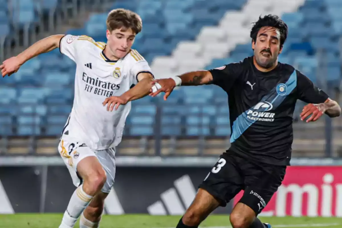 Two players compete for the ball during a match in a stadium with blue stands in the background. Two players compete for the ball during a match in a stadium with blue stands in the background.