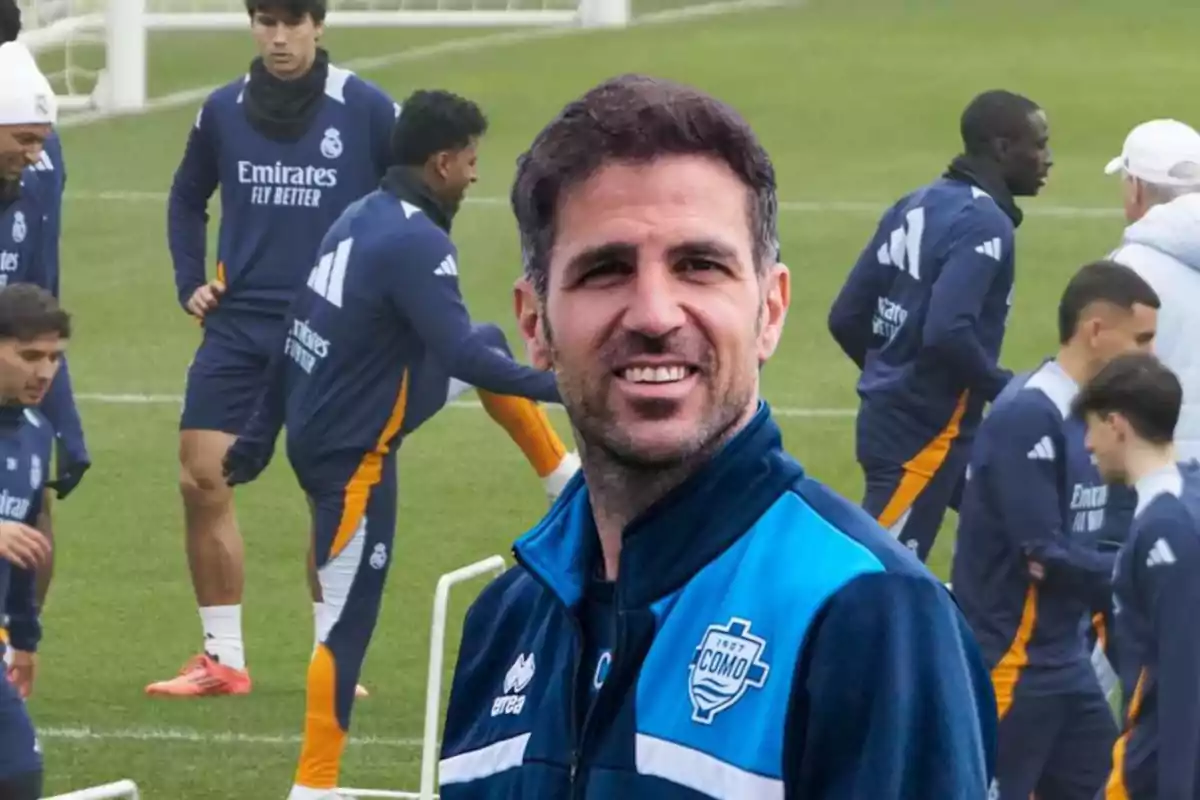 A smiling man in a blue soccer team jacket, with players training in the background.