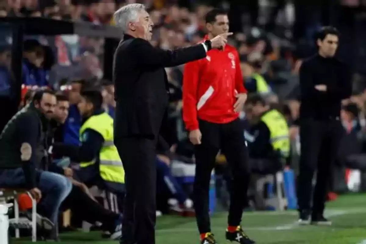 Un entrenador de fútbol da instrucciones desde la línea lateral durante un partido, mientras otros miembros del equipo técnico observan desde el banquillo.