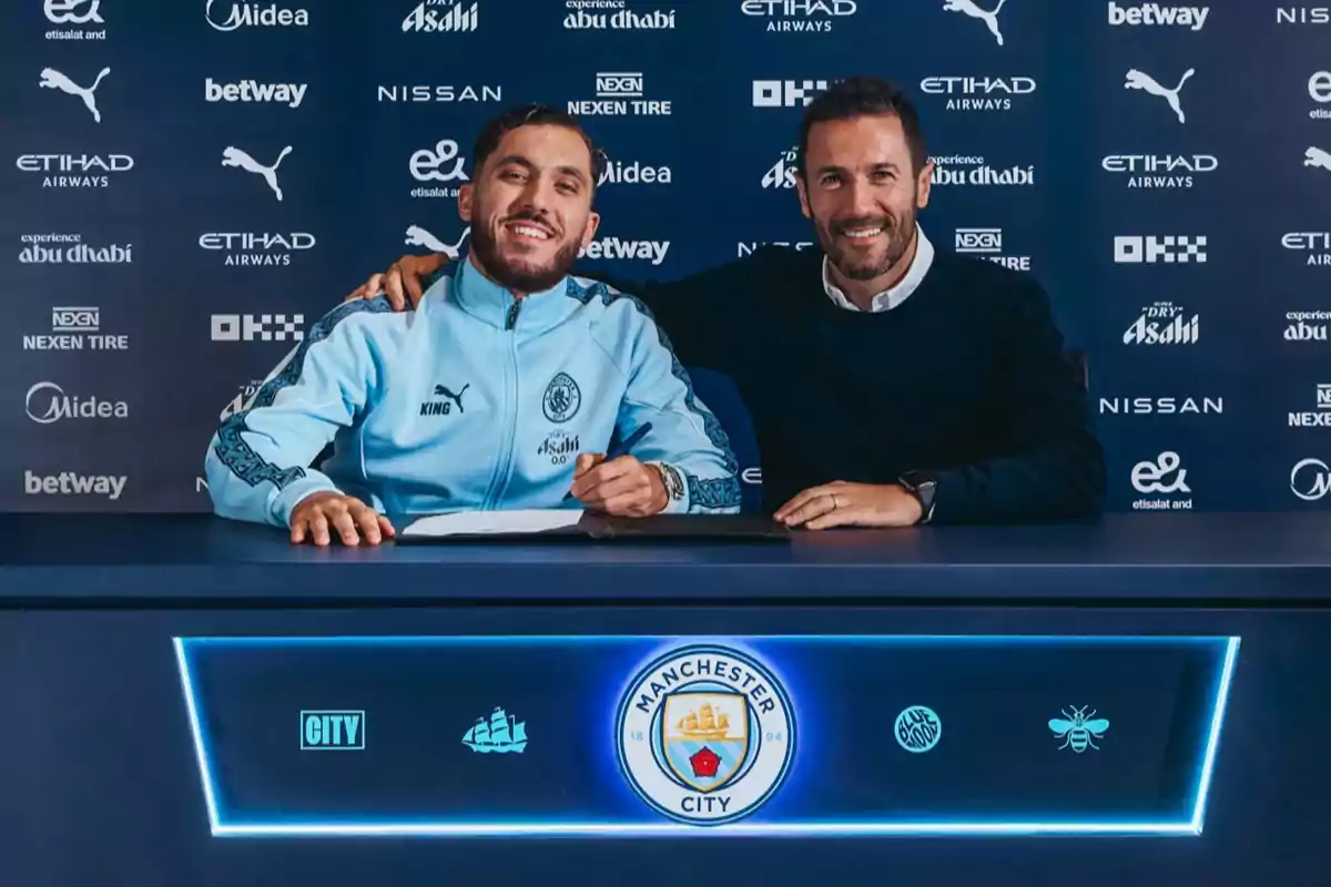 Dos hombres sonríen mientras uno de ellos firma un documento en una mesa con el logo del Manchester City al frente y un fondo azul con varios patrocinadores. Dos hombres sonríen mientras uno de ellos firma un documento en una mesa con el logo del Manchester City al frente y un fondo azul con varios patrocinadores.