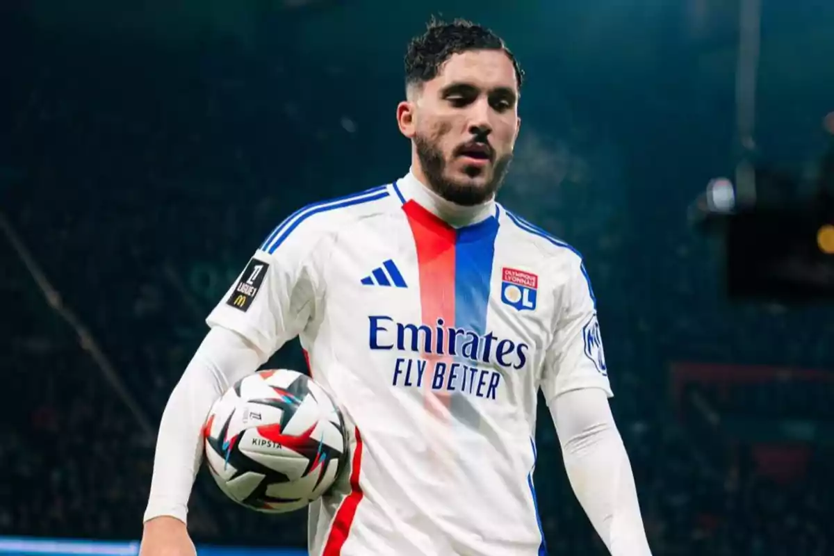 Jugador de fútbol con uniforme blanco y detalles en azul y rojo sosteniendo un balón en el campo. Jugador de fútbol con uniforme blanco y detalles en azul y rojo sosteniendo un balón en el campo.