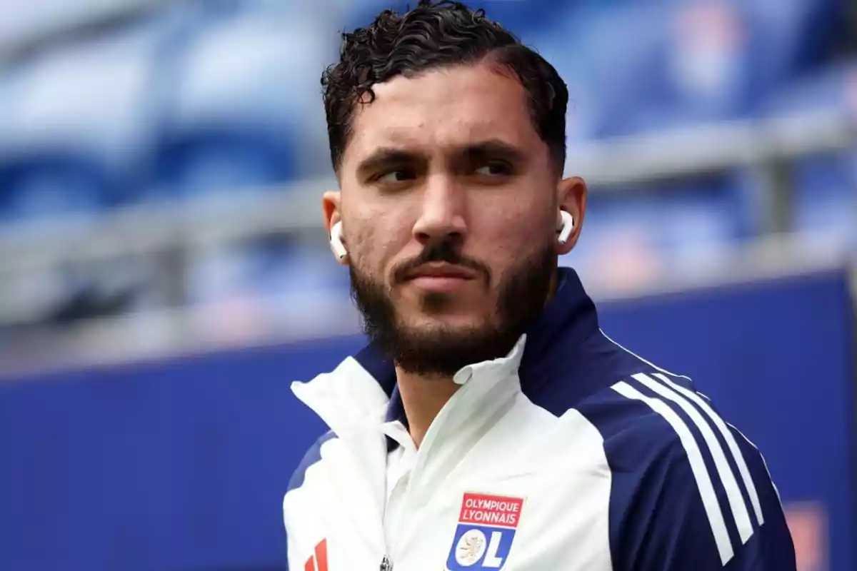 A man in a white and blue Olympique Lyonnais sports jacket, wearing headphones, looks to the side with a serious expression. A man in a white and blue Olympique Lyonnais sports jacket, wearing headphones, looks to the side with a serious expression.