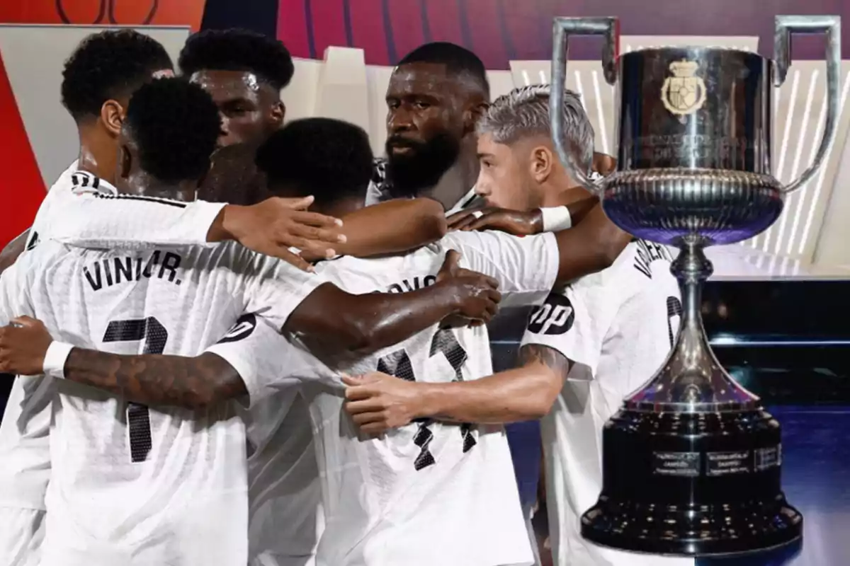 Jugadores de fútbol celebrando en grupo junto a un trofeo grande.