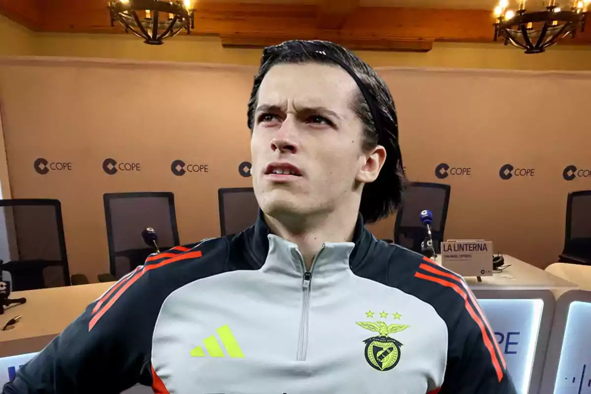 A player wearing a Benfica jacket is standing in front of a conference room with microphones and empty chairs.