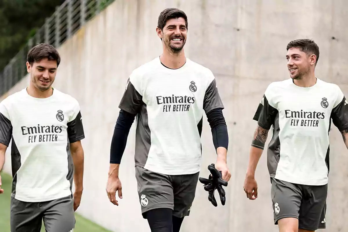 Three Real Madrid players smile as they walk together on a training field, wearing the team's sportswear. Three Real Madrid players smile as they walk together on a training field, wearing the team's sportswear.