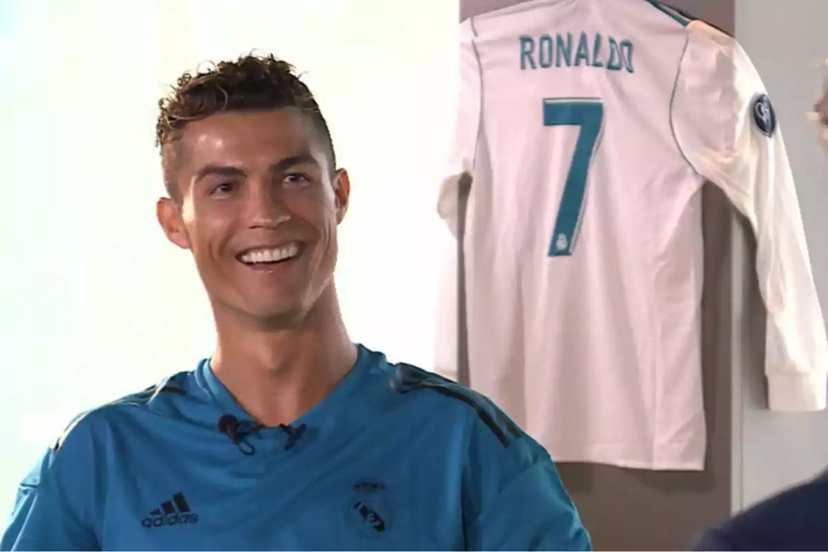 A smiling man in a blue soccer jersey with a white jersey hanging in the background.