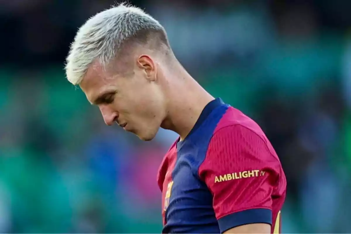A soccer player with blond hair and a serious expression wears a blue and red jersey on a playing field. A soccer player with blond hair and a serious expression wears a blue and red jersey on a playing field.