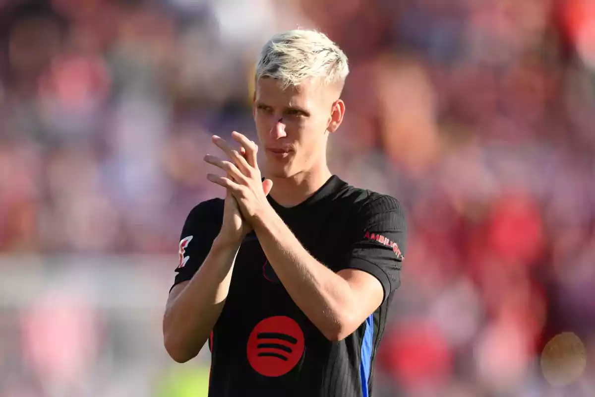 A soccer player with blonde hair applauds on the field while wearing a black jersey with a red logo. A soccer player with blonde hair applauds on the field while wearing a black jersey with a red logo.