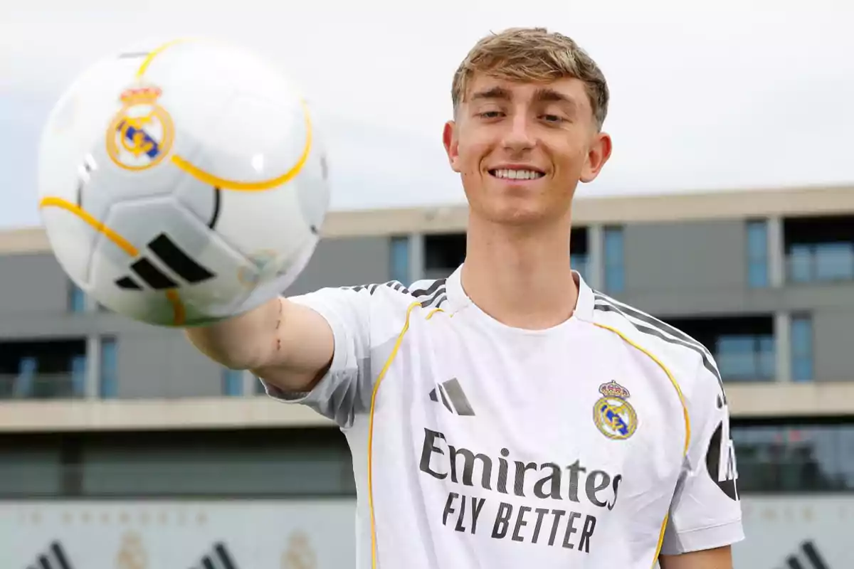Joven con camiseta del Real Madrid sosteniendo un balón de fútbol y sonriendo frente a un edificio moderno Joven con camiseta del Real Madrid sosteniendo un balón de fútbol y sonriendo frente a un edificio moderno