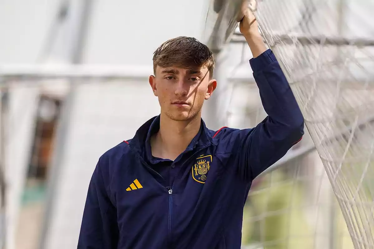 Un joven con chaqueta azul de la selección española de fútbol apoya su brazo en una portería. Un joven con chaqueta azul de la selección española de fútbol apoya su brazo en una portería.