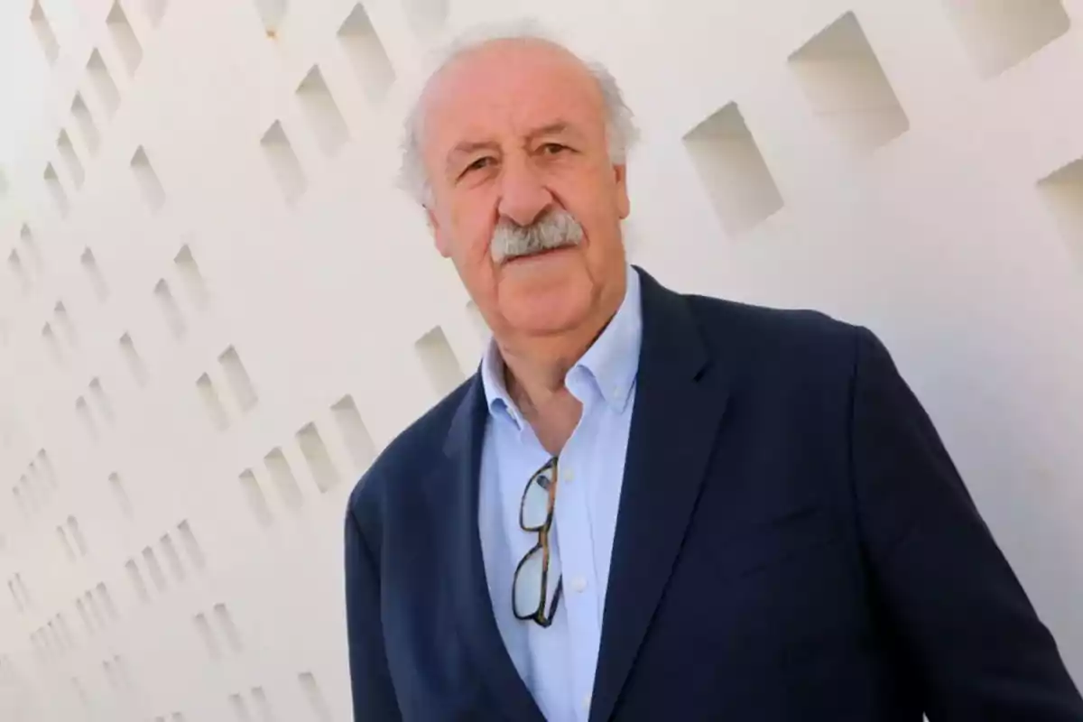 Un hombre mayor con bigote y cabello canoso viste un traje oscuro y camisa azul clara mientras posa frente a una pared blanca con un patrón geométrico.