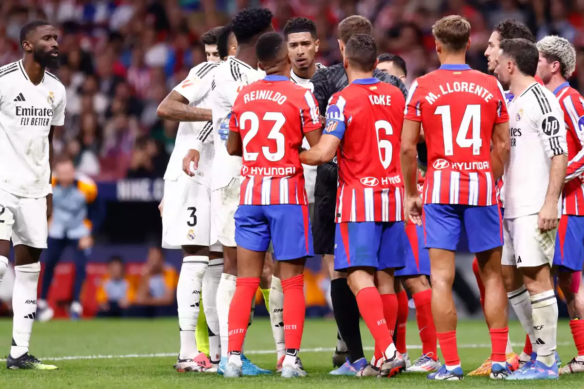 Jugadores de dos equipos de fútbol, uno con uniforme blanco y otro con uniforme rojo y azul, discuten en el campo durante un partido.