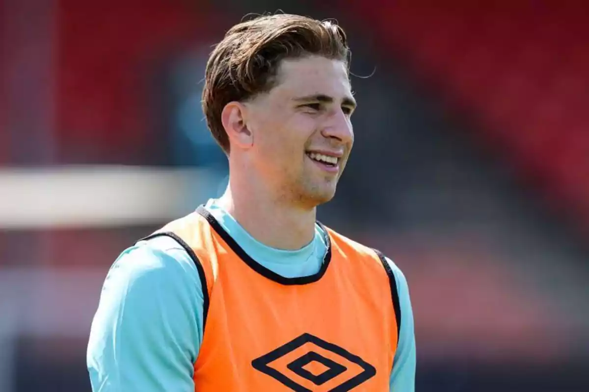 A soccer player smiles while wearing an orange training bib on a sports field.