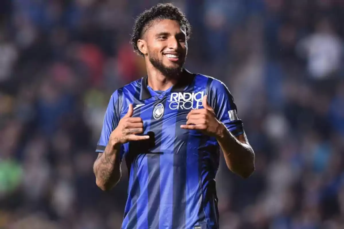 A soccer player smiles while gesturing with his hands wearing a blue and black uniform in a stadium.