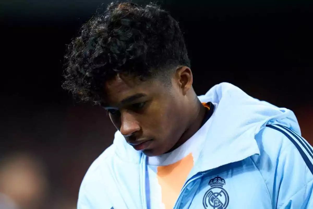 A young man in a light blue sports jacket with a soccer team logo, looking down with a thoughtful expression. A young man in a light blue sports jacket with a soccer team logo, looking down with a thoughtful expression.