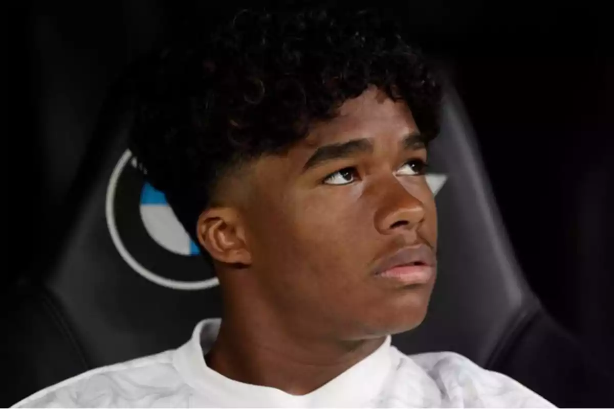 Un joven con cabello rizado y expresión pensativa mira hacia arriba, sentado en un asiento con un logotipo en el fondo. Un joven con cabello rizado y expresión pensativa mira hacia arriba, sentado en un asiento con un logotipo en el fondo.
